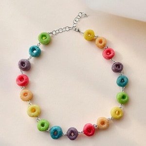 Peut inclure: Un collier composé d'une série de petites perles en forme d'anneau de différentes couleurs, notamment rouge, orange, jaune, vert, bleu et violet. Les perles sont reliées par une chaîne en métal argenté avec un fermoir mousqueton et une chaînette.