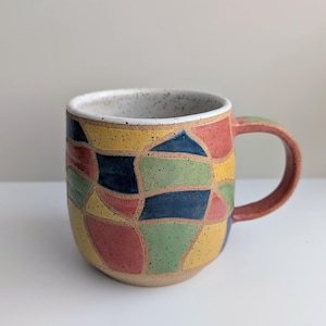 May include: A ceramic mug featuring a speckled off-white interior and a multi-colored exterior pattern of irregular, hand-painted shapes in red, yellow, blue, and green. The mug has a rounded body, a sturdy handle, and a speckled clay base.