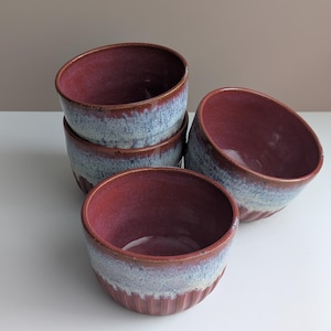 May include: A set of four ceramic bowls featuring a deep reddish-maroon interior and a reactive glaze finish. The exterior displays a gradient transition from a dark maroon rim to a textured, light blue and white speckled base with vertical ribbing.