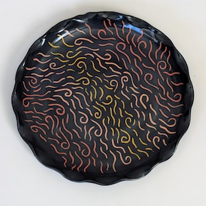 May include: A round black ceramic plate with a scalloped edge, featuring a hand-painted pattern of small, wavy, and swirling lines in shades of yellow, orange, and pink scattered across the entire surface of the dish.