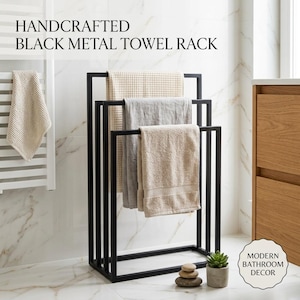 Peut inclure: Un porte-serviettes en métal noir avec un cadre rectangulaire, doté de trois barres étagées supportant une serviette beige gaufrée, une serviette gris clair et une serviette beige. Le texte indique: HANDCRAFTED BLACK METAL TOWEL RACK et MODERN BATHROOM DECOR.