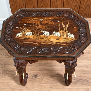 May include: An octagonal wooden side table with a dark brown finish. The tabletop features an inlaid scene of five white elephants walking among trees and birds. The table legs are carved into the shape of elephant heads with white tusks.