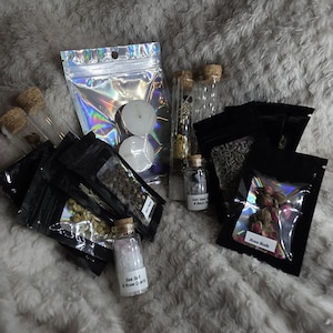May include: A collection of small glass bottles with cork stoppers, small black pouches, and a holographic bag. The bottles contain herbs and crystals. The pouches contain dried herbs and rose buds. The labels read "Sea Salt & Rose Quartz" and "Dark Black & Black Salt".