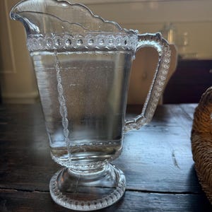 May include: A clear glass pitcher with a decorative rim and handle. The pitcher has a scalloped edge and a detailed design around the base and handle. The pitcher is filled with a clear liquid, likely water, and sits on a dark wooden surface.
