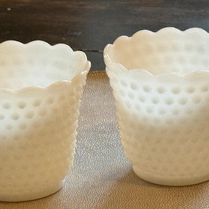 May include: Two off-white, hobnail-textured glass vases with scalloped rims. The vases are cylindrical with a slightly flared base. They sit on a gold-toned surface, and the background is a dark wooden table.
