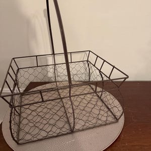 May include: A rustic, rectangular wire basket with a handle, perfect for storage or display. The basket is made of dark brown metal with a chicken wire mesh design. It sits on a round, textured, light-colored placemat.