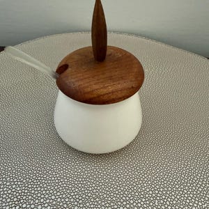 May include: A white ceramic sugar bowl with a wooden lid and handle. A small, clear spoon rests on the side. The lid is a rich brown color, and the handle is a pointed, conical shape. The bowl sits on a textured, light gray surface.