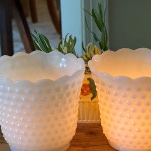May include: Two white hobnail glass candle holders with scalloped rims. One is lit, emitting a warm glow. The hobnail texture adds visual interest. The candle holders are likely used for decorative purposes.