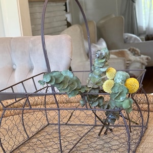May include: A rectangular wire basket with a tall arched handle, constructed from dark brown metal mesh. Inside the basket are several stems of dried green eucalyptus leaves and three bright yellow, spherical billy ball flowers.