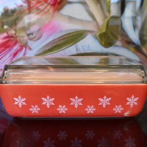 May include: A rectangular, orange Pyrex baking dish with a clear glass lid. The dish is decorated with white snowflake patterns. A vintage kitchenware item, perfect for baking and serving.