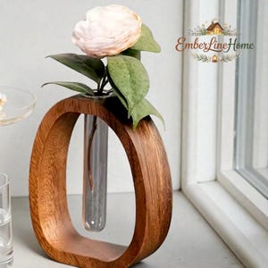Può includere: Un vaso di legno con una cornice ovale, che contiene un tubo di vetro trasparente con un fiore rosa chiaro e foglie verdi. Il vaso è realizzato in legno dai toni caldi e si trova su un davanzale. Il logo "EmberLineHome" è visibile.