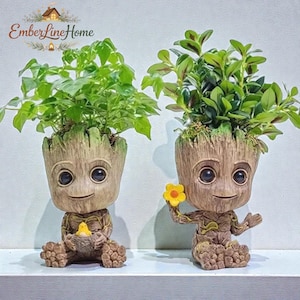 Puede incluir: Dos macetas con forma de Groot, cada una con una planta verde vibrante. Una sostiene un pajarito amarillo, la otra una flor amarilla. Las macetas son marrones con texturas detalladas de madera. El texto "EmberLineHome" está en la parte superior.