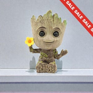 May include: A Groot figurine, a character from Guardians of the Galaxy, holding a yellow flower. The figure is brown with green detailing and has large, black eyes. The figurine is on a white shelf.