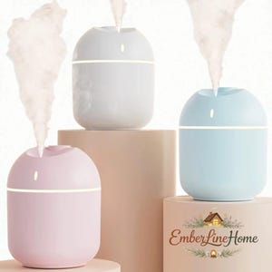 Op de afbeelding: Drie pastelkleurige etherische olie diffusers. De diffusers zijn lichtroze, lichtblauw en wit, elk met een gloeiende horizontale band. Elke diffuser geeft een stroom stoom af. Het EmberLineHome-logo is rechtsonder zichtbaar.