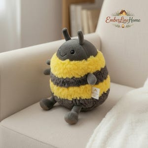 Puede incluir: Un juguete de peluche de abeja con una cara sonriente, antenas grises y pequeños brazos y piernas grises. La abeja tiene pelaje a rayas amarillas y grises. Una pequeña etiqueta está adherida al cuerpo de la abeja.