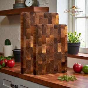 May include: Two wooden chopping boards with a mosaic pattern of rectangular blocks. The boards are various shades of brown and sit on a wooden countertop. The boards are next to a window with a plant and some apples.