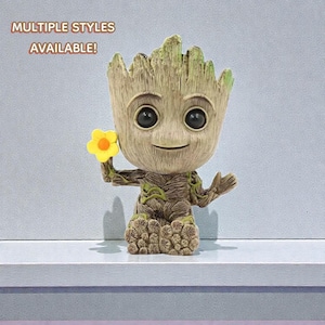 May include: A Groot figurine with a yellow flower. The figurine is a light brown color with dark brown and green accents. The text "MULTIPLE STYLES AVAILABLE!" is in the upper left corner. The figurine is on a white shelf.