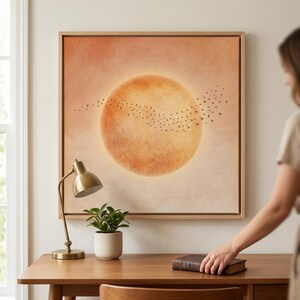 Könnte beinhalten: Gerahmtes Wandbild mit einer großen orangefarbenen Sonne und einem Vogelschwarm. Das Kunstwerk hängt über einem Holzschreibtisch mit einer Messinglampe und einer kleinen Topfpflanze. Warme Farbpalette.