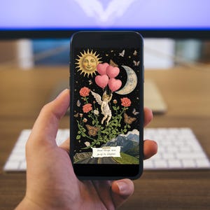 Hand holding a phone showing a mixed media collage wallpaper with celestial and botanical art in a cozy room setting