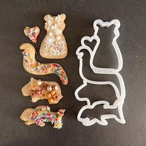 Peut inclure: Plusieurs biscuits décorés de formes variées, dont un cœur, une robe, un ver, un animal en forme d'étoile et un poisson. Les biscuits sont ornés de vermicelles colorés et de décorations comestibles. Un emporte-pièce en plastique blanc avec le mot "Teigtige" est également présent.