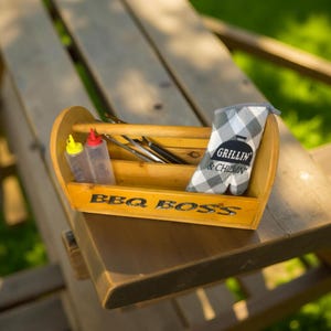May include: Wooden BBQ caddy with the text "BBQ BOSS" and a handle. It contains two squeeze bottles, grilling tools, and a checkered oven mitt that says "Grillin' & Chillin'". The caddy is sitting on a wooden picnic table.