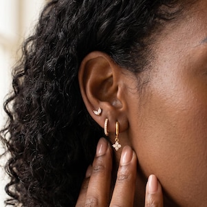 May include: A close-up of an ear displaying three gold earrings. The earrings include a small crescent moon stud, a small hoop with embedded crystals, and a dangling four-petal flower design. The earrings are gold in colour.