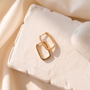 May include: A pair of gold-toned hoop earrings featuring a rectangular shape with rounded corners. The earrings have a polished metallic finish and a hinged closure mechanism, designed as a continuous loop that forms a distinct oblong silhouette.