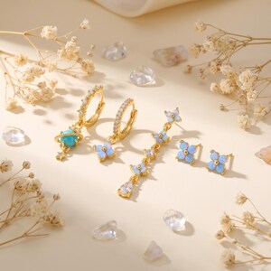 May include: A set of gold-tone earrings featuring a hoop with a turquoise planet and star charm, a hoop with a four-petal light blue flower, a long drop earring with multiple flower charms, and two small four-petal light blue flower stud earrings.