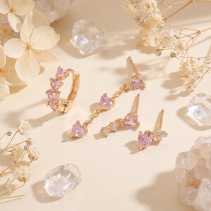 May include: A set of gold-tone earrings featuring pink heart-shaped stones. The collection includes a hinged hoop earring with a row of hearts, a dangling chain earring with heart accents, and two stud earrings with heart and clear stone details.