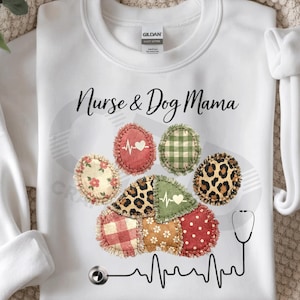 Puede incluir: Sudadera blanca con el texto "Nurse & Dog Mama" sobre un diseño de huella de pata colorido. La huella de la pata está hecha de parches con estampados florales, a cuadros y de leopardo. Un estetoscopio y un gráfico de latido cardíaco completan el diseño.