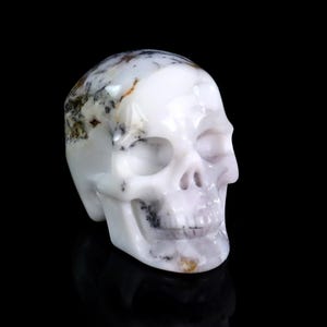 May include: A carved skull figurine in white with gray and brown inclusions. The skull has detailed features, including eye sockets, a nose cavity, and teeth. The polished surface reflects light, set against a black background.