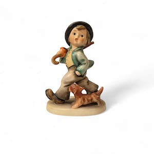 May include: A ceramic figurine of a young boy walking with a dog. The boy wears a green jacket, brown pants, and a black hat. He carries a walking stick and an umbrella. The dog is brown and tan. The figurine is on a beige base.