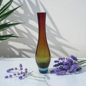 May include: A tall, slender glass vase with a gradient of colors, transitioning from a deep red at the top to amber and ending with a light blue band at the base. The vase is displayed with sprigs of purple lavender.