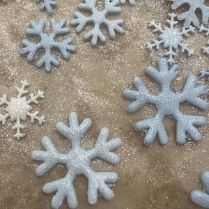 May include: An assortment of snowflake decorations in varying sizes and shades of white and light blue. The snowflakes are arranged on a brown surface, some with a glittery finish, evoking a winter or festive theme.