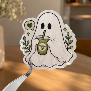 May include: A die-cut sticker featuring a white ghost character holding a green matcha drink in a clear plastic cup with a straw. The cup has the text "matcha club" printed on it. The design includes small green leaves, a heart, and stars.