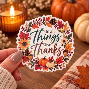 May include: A circular sticker featuring a decorative autumn wreath of colourful leaves, berries, pinecones, sunflowers, and pumpkins surrounding the text In all Things Give Thanks. The design includes holographic accents that catch the light.