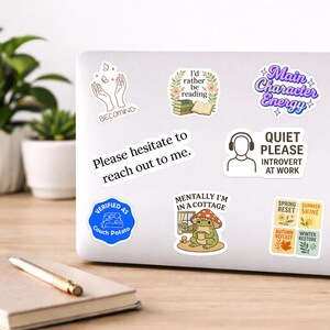 May include: A collection of colorful stickers on a laptop. The stickers feature various designs, including text like "Main Character Energy," "Quiet Please Introvert at Work," and a frog in a cottage. Other stickers have phrases like "Verified as Couch Potato."