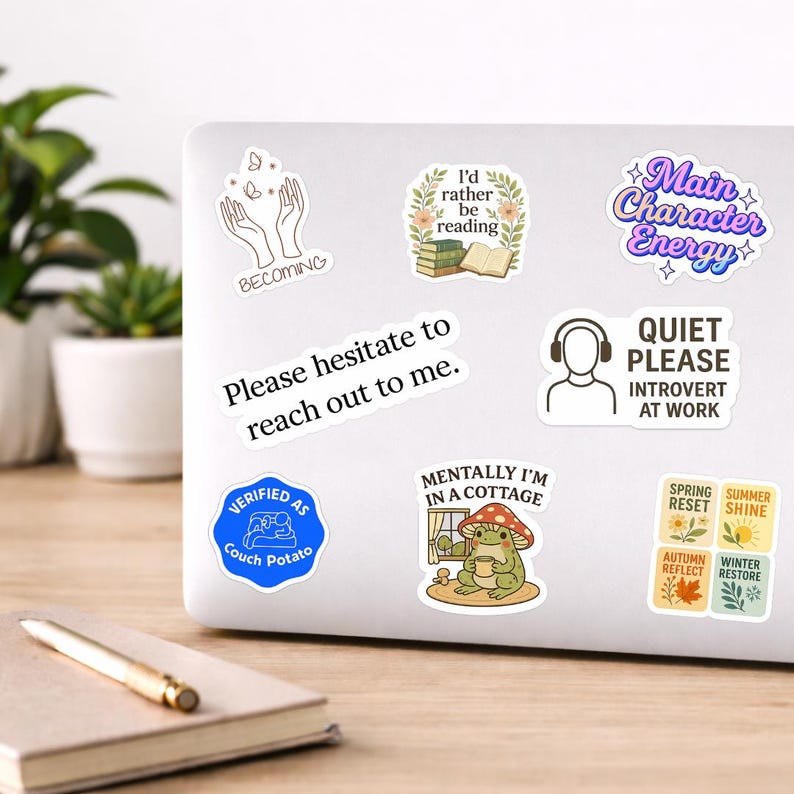 May include: A collection of colorful stickers on a laptop. The stickers feature various designs, including text like "Main Character Energy," "Quiet Please Introvert at Work," and a frog in a cottage. Other stickers have phrases like "Verified as Couch Potato."