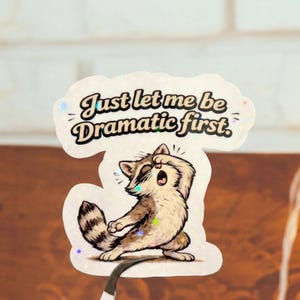 May include: A sticker featuring a raccoon in a dramatic pose, with the text "Just let me be Dramatic first." The raccoon is light brown and white with a black striped tail. The sticker has a holographic, glittery finish.