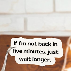 May include: A white sticker with the text "If I'm not back in five minutes, just wait longer." The sticker is held by a pair of silver tweezers against a brown surface and a white brick wall background.