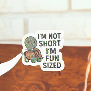 May include: A glittery sticker featuring a cartoon turtle with a brown shell and green limbs. The sticker has the text "I'M NOT SHORT I'M FUN SIZED" in black capital letters. The sticker is held by silver tweezers.