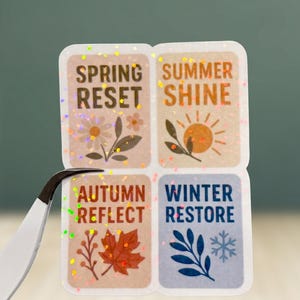 May include: A set of four seasonal stickers with glitter accents. Each sticker features a season's name and a related image: Spring Reset, Summer Shine, Autumn Reflect, and Winter Restore. A pair of tweezers holds the stickers.