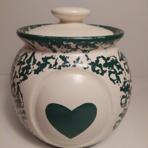 Peut inclure: Un pot en céramique blanc cassé de forme ronde avec un couvercle assorti. Le pot présente un cœur vert foncé centré sur le devant et un motif éponge vert foncé autour du bord et de la partie supérieure. La surface en céramique est mate.