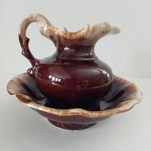 Peut inclure: Un ensemble vintage de pichet et de bassin en céramique marron. Le pichet a une anse incurvée et un bord évasé avec un bord de couleur crème. Le bassin a un bord de couleur crème assorti et un motif festonné. L'ensemble est brillant et reflète la lumière.