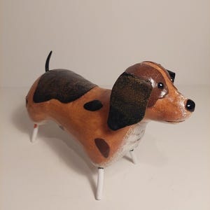 May include: A decorative dog figurine featuring a brown and tan body with dark brown spots, black ears, and a small black nose. The sculpture stands on four thin, white metal legs and has a short, upright tail, showcasing a smooth finish.