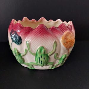May include: A ceramic planter with a lotus flower design. The planter is white with red and pink accents. Three green frog figurines are attached to the planter. The planter has a scalloped edge and is on a black surface.