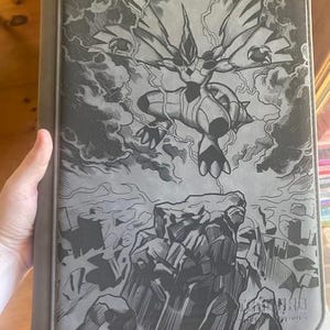 May include: A grey trading card binder featuring a black and grey graphic illustration of a Zekrom creature flying above a rocky landscape with lightning bolts in the background. The bottom right corner displays the text Trading Cards.