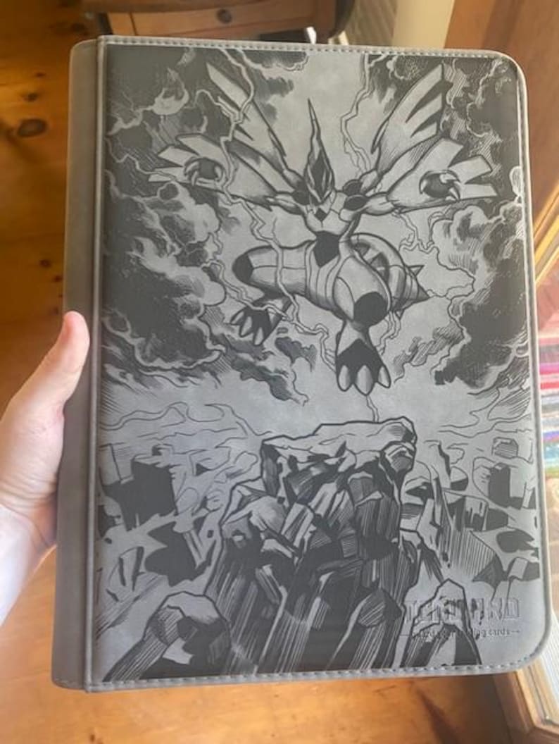 May include: A grey trading card binder featuring a black and grey graphic illustration of a Zekrom creature flying above a rocky landscape with lightning bolts in the background. The bottom right corner displays the text Trading Cards.