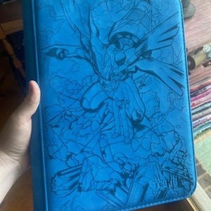May include: A bright blue rectangular binder featuring a dark blue line art illustration of a large mechanical creature and a human figure standing amidst rubble. The design covers the entire front cover, which has a zippered edge.