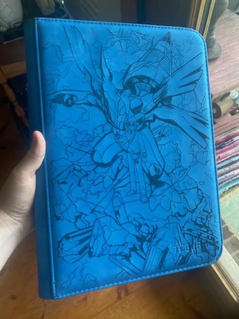 May include: A bright blue rectangular binder featuring a dark blue line art illustration of a large mechanical creature and a human figure standing amidst rubble. The design covers the entire front cover, which has a zippered edge.